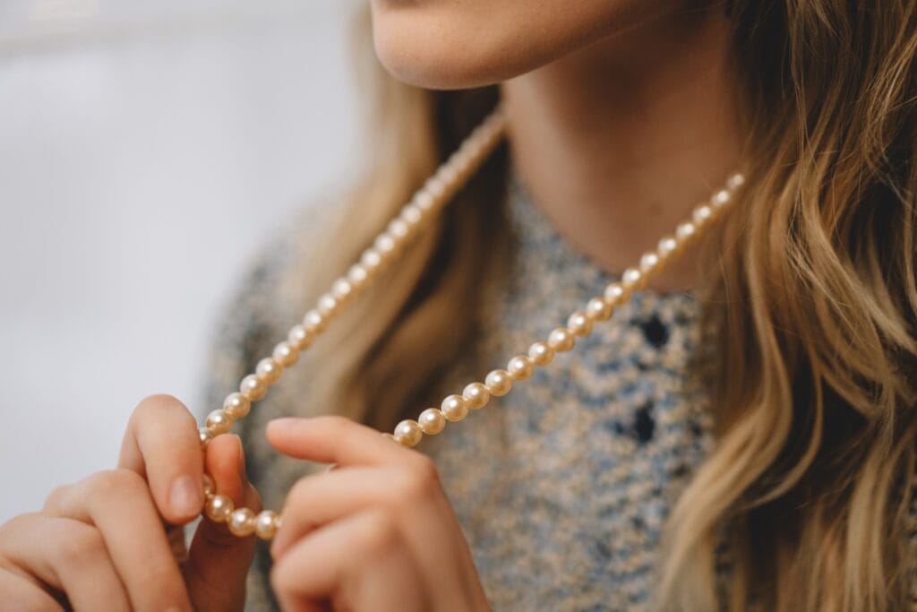 A fashionable woman holding a pearl necklace with blonde hair visible in a stylish close-up shot.