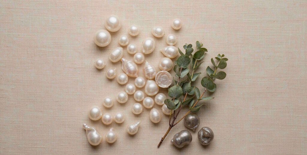 Assorted natural freshwater pearls in different shapes — round, button, drop, and baroque — alongside fresh eucalyptus sprigs on beige fabric. A cover image celebrating the organic diversity of real freshwater pearls from Zhuji, China.
